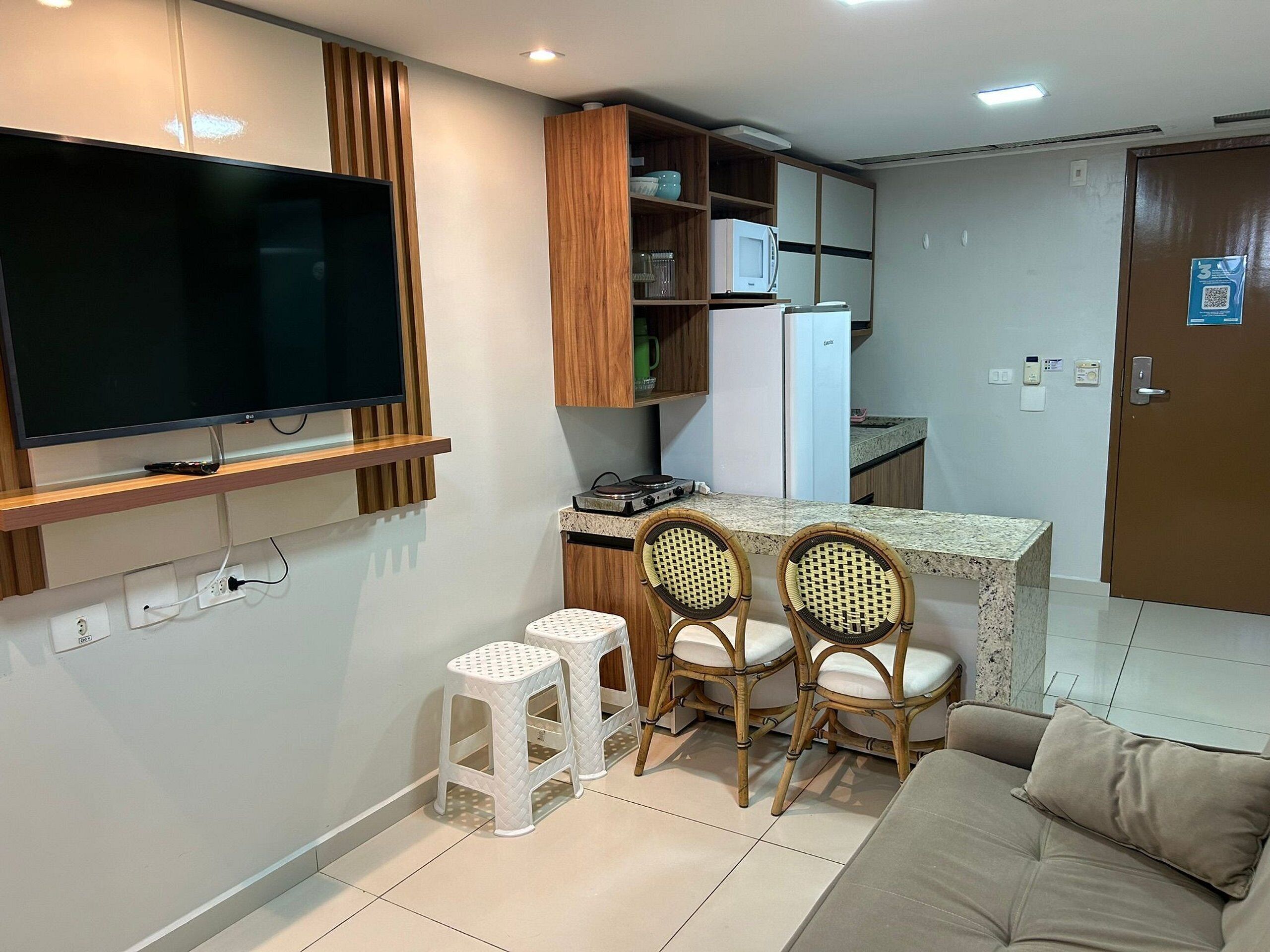 A modern kitchen with a flat-screen TV, wooden cabinets, a microwave, and a refrigerator. There are two wicker chairs and a small white stool.