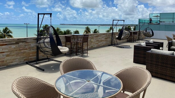 A rooftop terrace with wicker furniture, a glass table, and a view of the ocean. - BELA HOSPEDAGEM-Imperial Flats em Tambaú (João Pessoa)