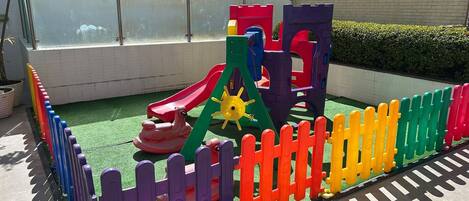 Un espace de jeux pour enfants avec un toboggan, des structures d’escalade et une zone de jeux délimitée par une clôture.
