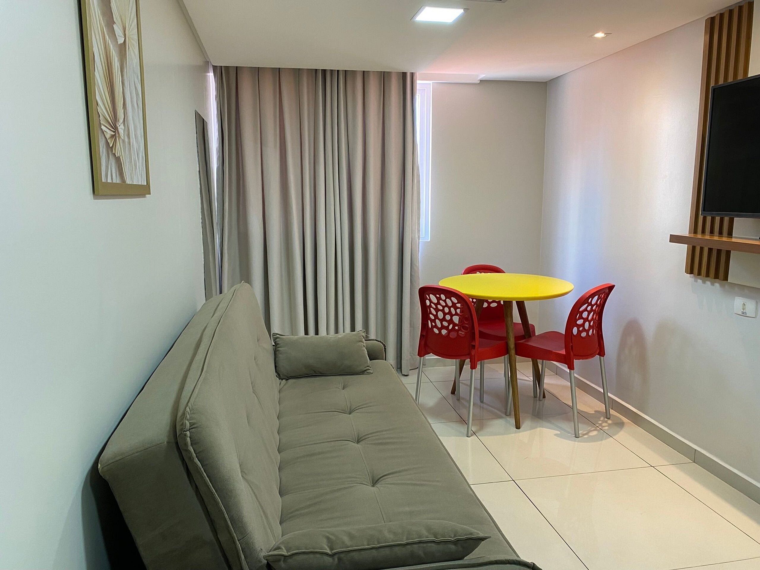 A modern living room with a sofa, a small dining table with chairs, a TV mounted on the wall, and a window with curtains.