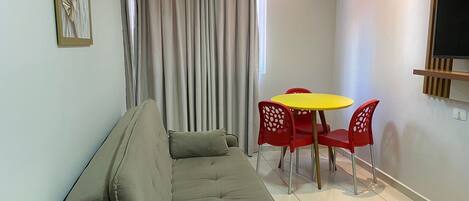 A modern living room with a sofa, a small dining table with chairs, a TV mounted on the wall, and a window with curtains.