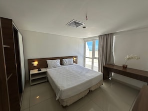A hotel room with a bed, a nightstand, a desk, a window with curtains, and a view of the outdoors.