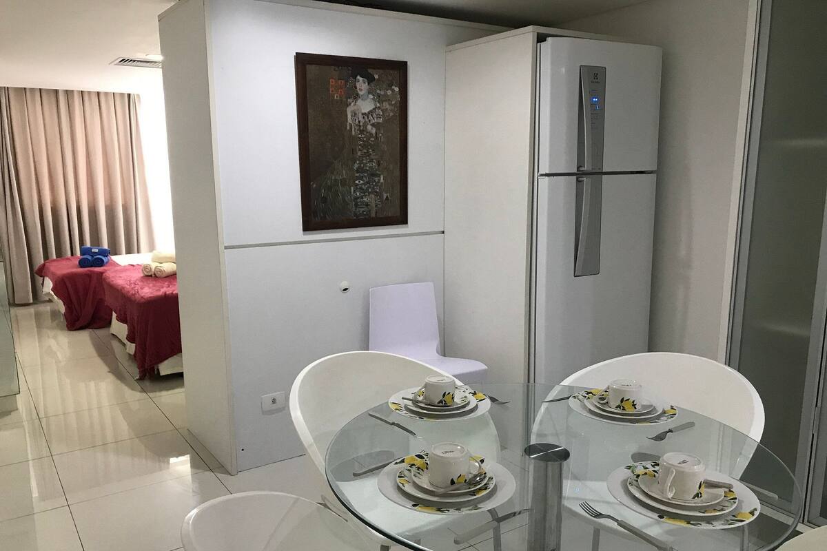 Un espacio habitable compacto con comedor que incluye una mesa de vidrio dispuesta para dos personas, una silla blanca y un refrigerador.