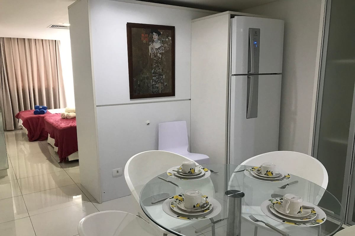 Une chambre dâhĂŽtel moderne avec une table Ă manger en verre, mise pour deux personnes, une chaise blanche, une petite table blanche, une table rouge avec des serviettes, un tableau au mur et un rĂ©frigĂ©rateur blanc.
