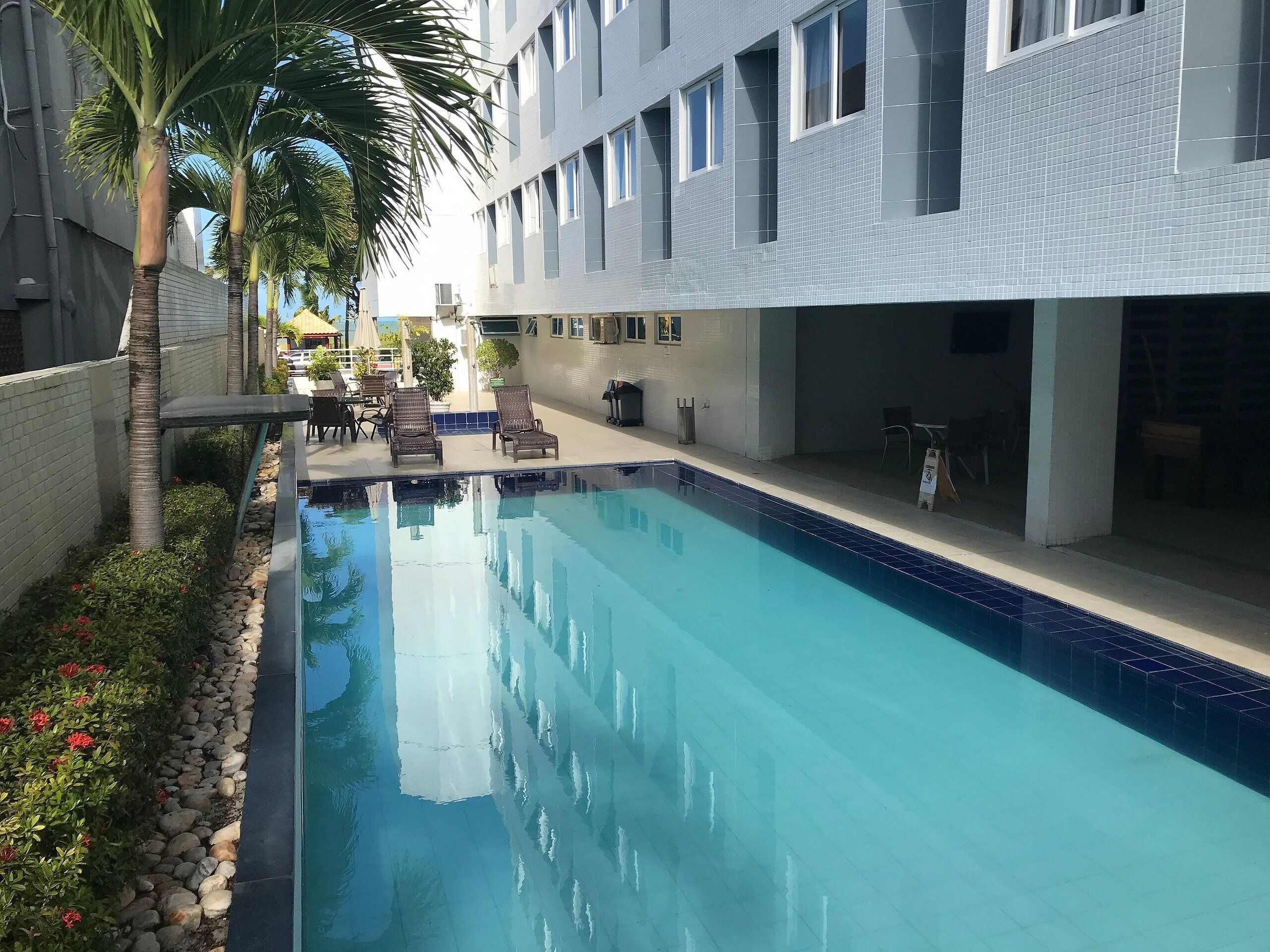 En swimmingpool med liggestole, en palme og en bygning med mange vinduer.