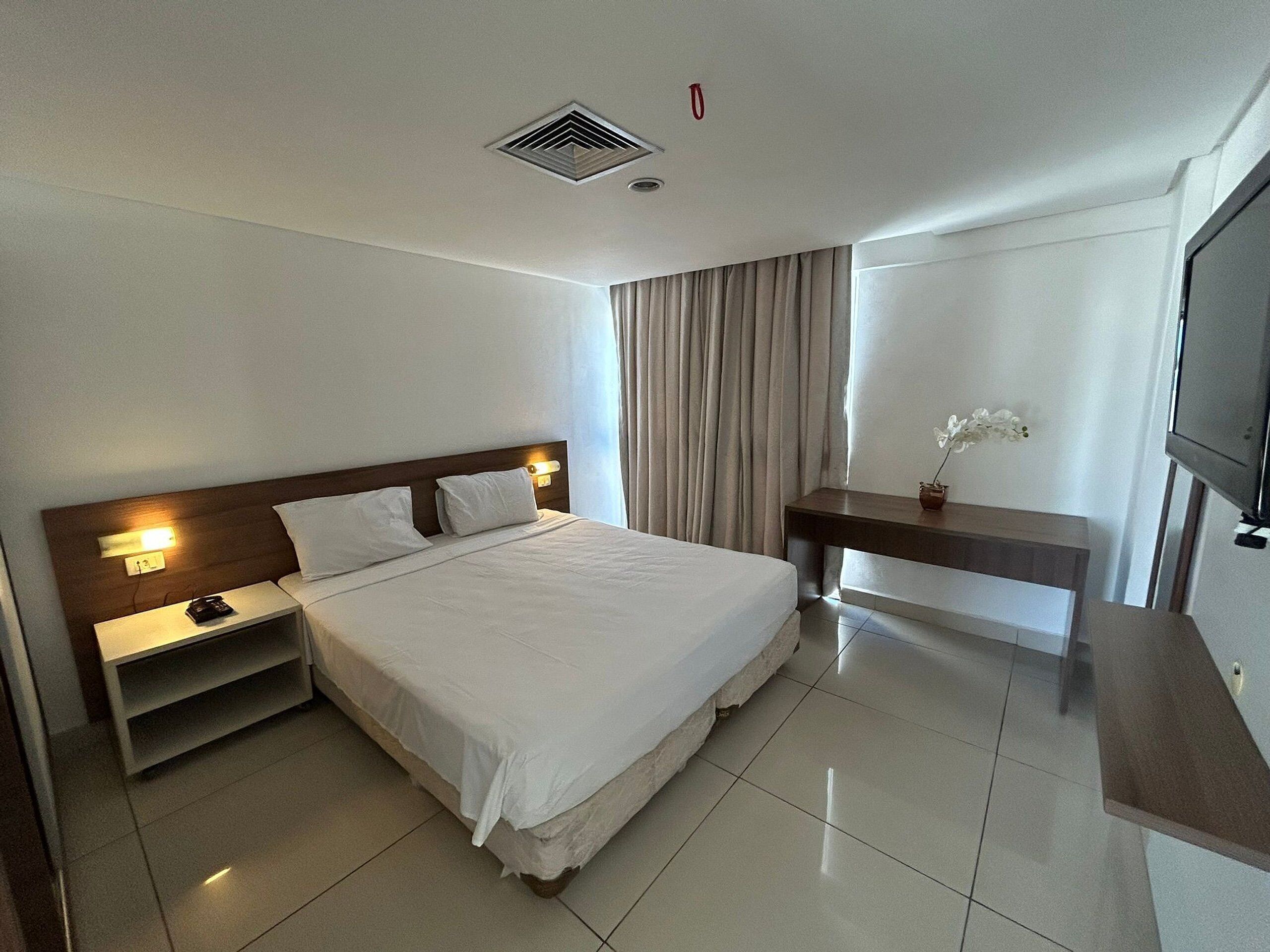 A hotel room with a bed, bedside table, lamp, TV, and curtains.