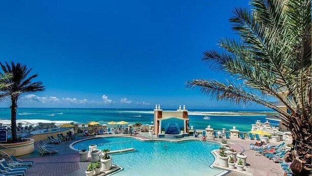 A resort pool area with a view of the ocean, lounge chairs, and umbrellas. - Bright Corner Unit with Stunning Gulf Views & Sunsets (Destin)