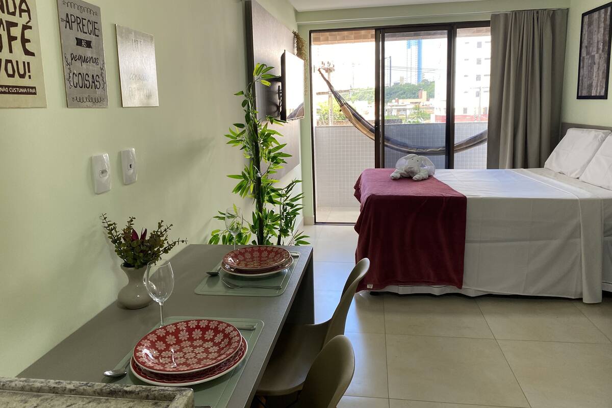 Habitación de hotel con cama, una mesa de comedor con platos y un vaso, una planta en maceta y vistas de edificios a través de la ventana.