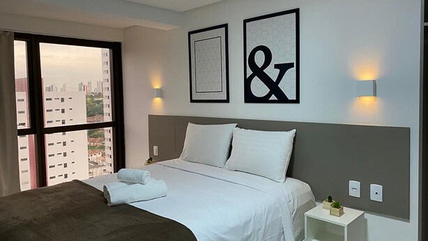 Hotelkamer met een groot bed, nachtkastje, klein tafeltje met een plant en uitzicht op de stad door een groot raam.