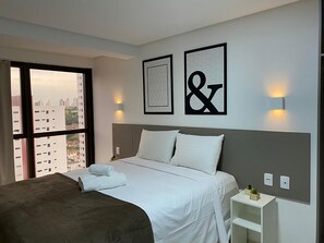 Ett hotellrum med en stor säng, ett nattduksbord, ett litet bord med en växt och utsikt över staden genom ett stort fönster.