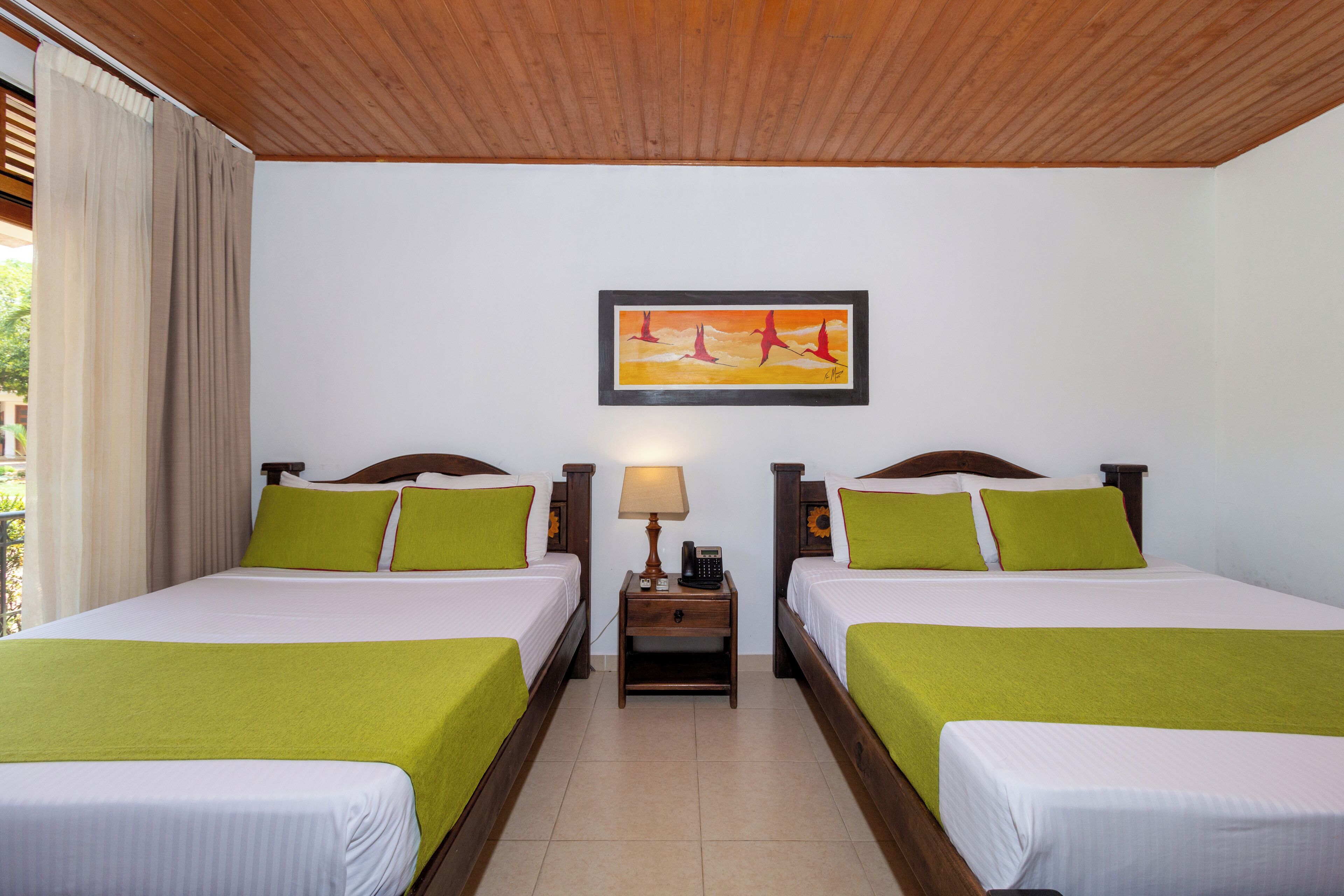 A hotel room with two beds, each with green bedding, a nightstand with a lamp, and a framed painting above the beds.