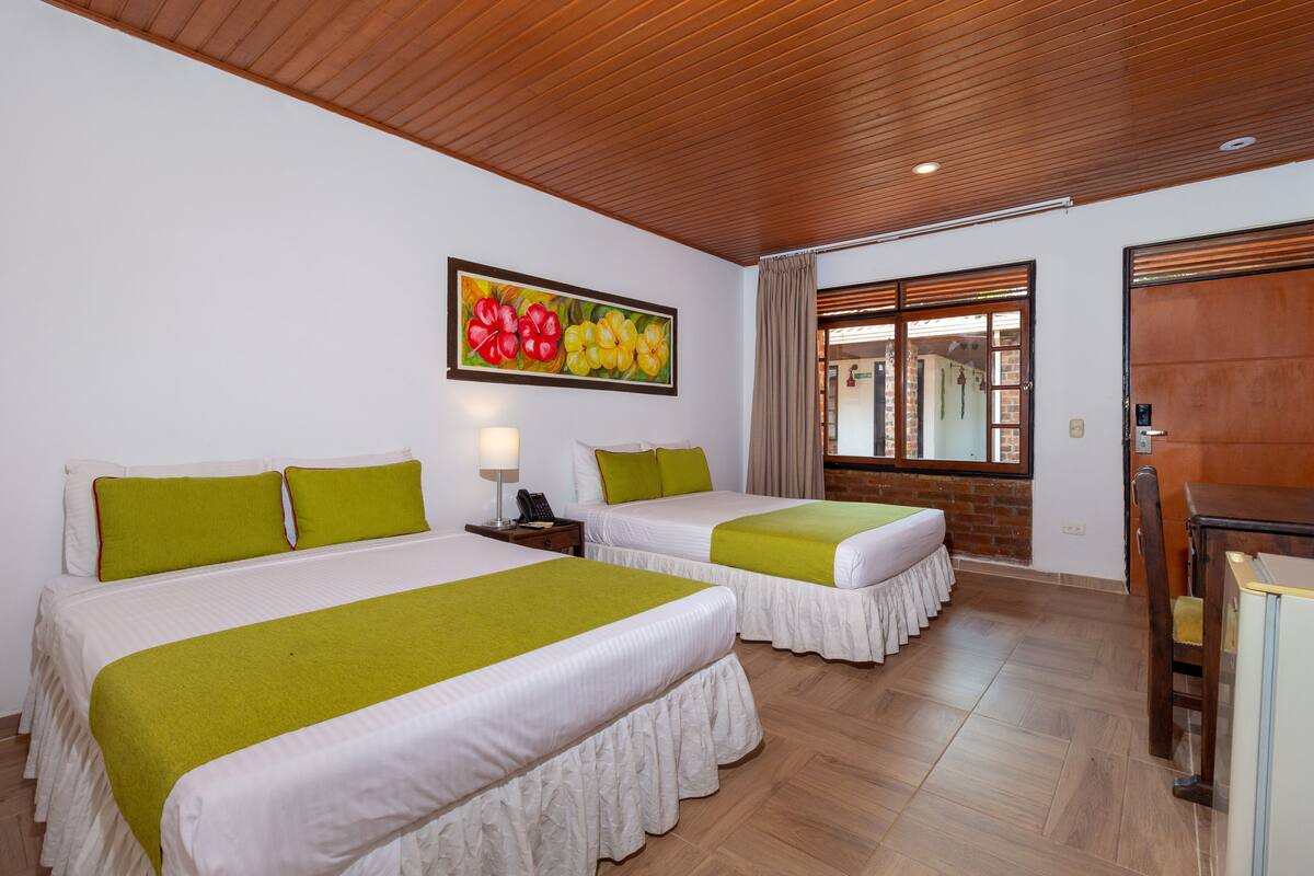 A hotel room with two beds, a painting of flowers on the wall, a wooden ceiling, and a window with curtains.