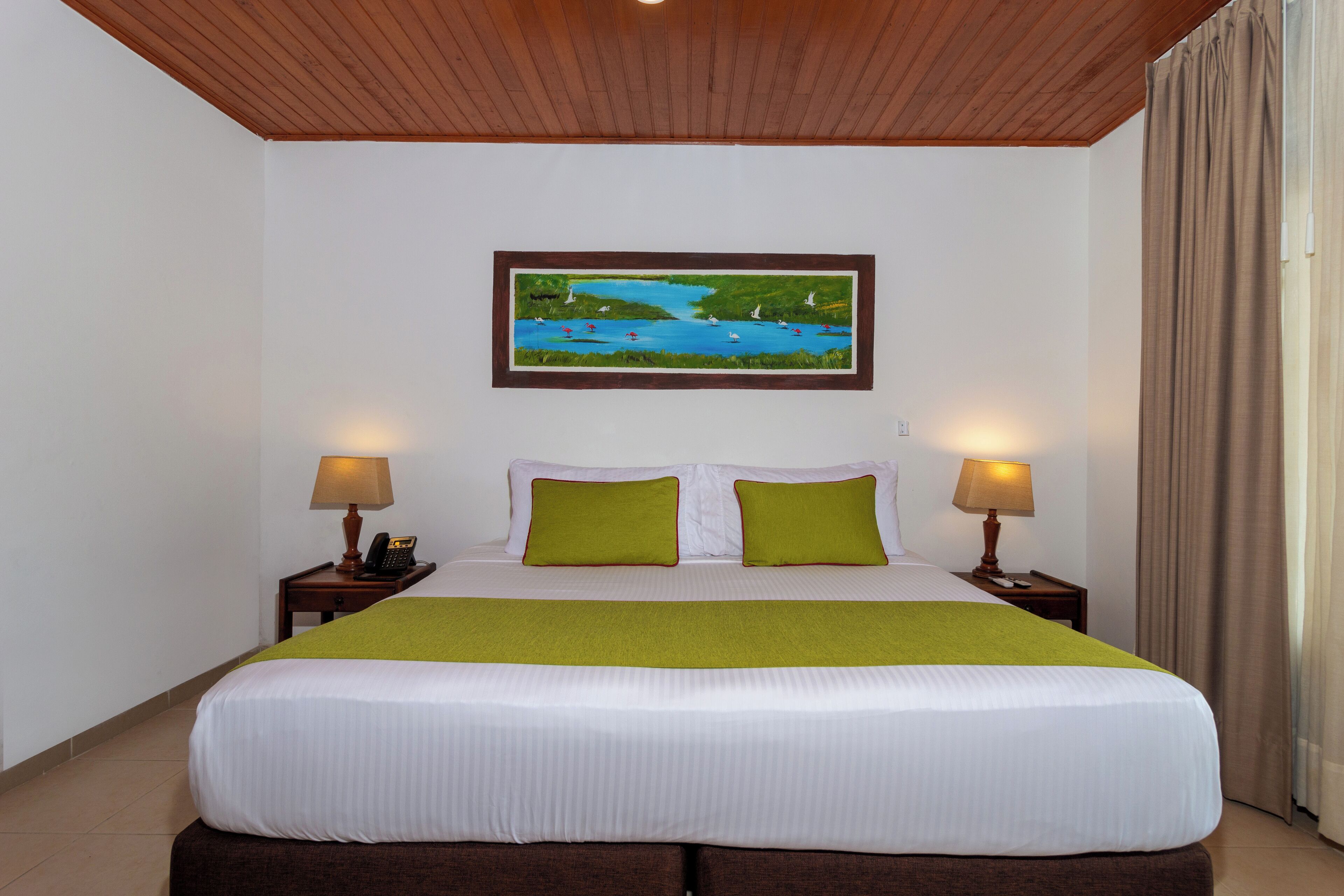 Una habitación de hotel con una cama grande, dos lámparas de noche, un cuadro de un lago y una ventana con cortinas.