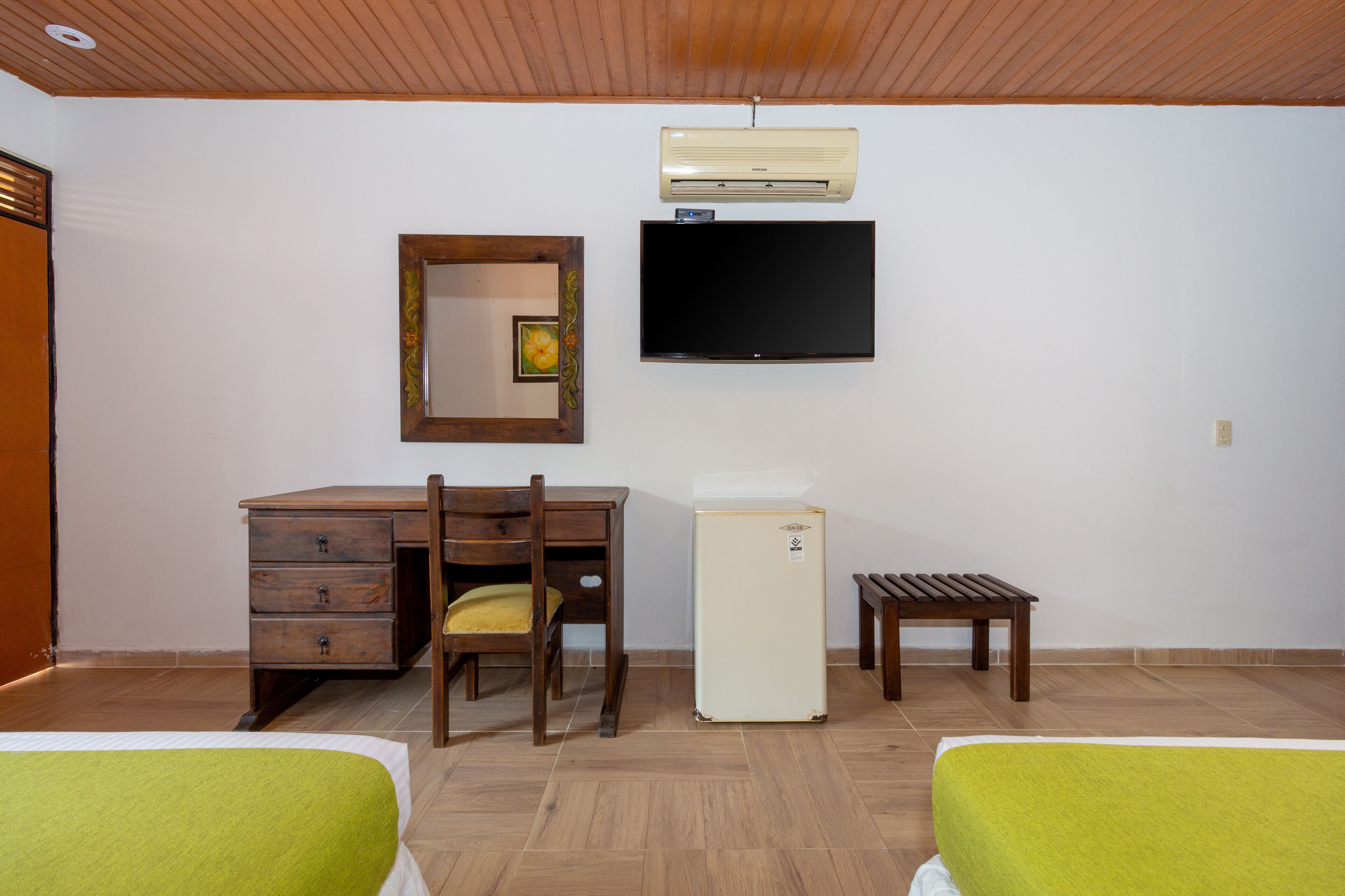 A room with a wooden desk, a chair, a small refrigerator, a television, and two beds with green bedding.