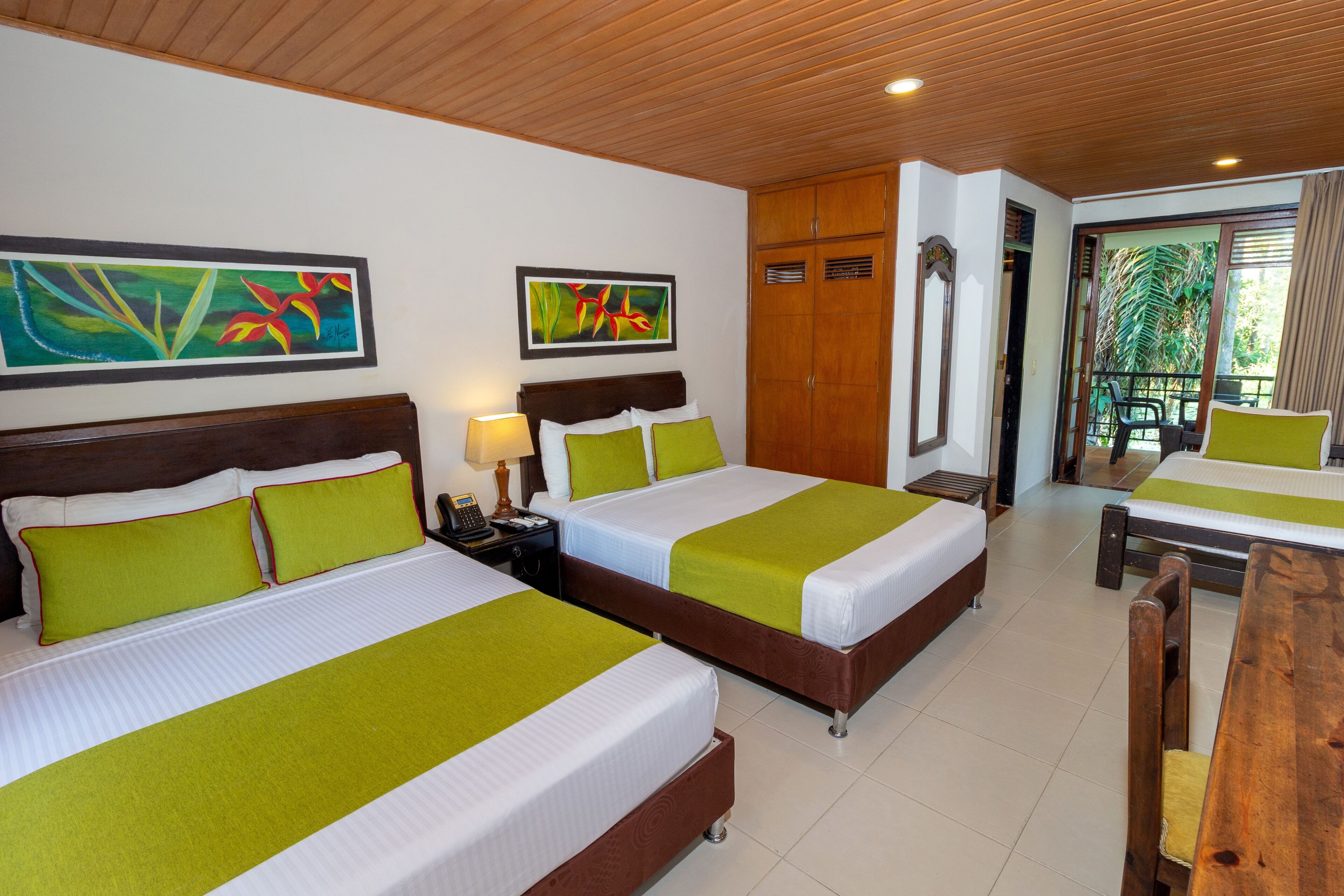 A hotel room with two beds, a wooden headboard, green pillows, a nightstand, a desk, and a view of the outdoors.
