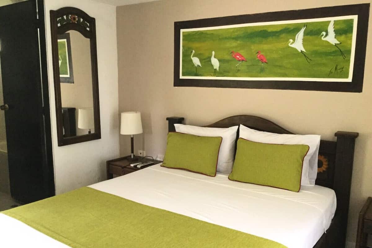 A hotel room with a bed, green pillows, a nightstand with a lamp, a framed painting of birds, and a mirror.