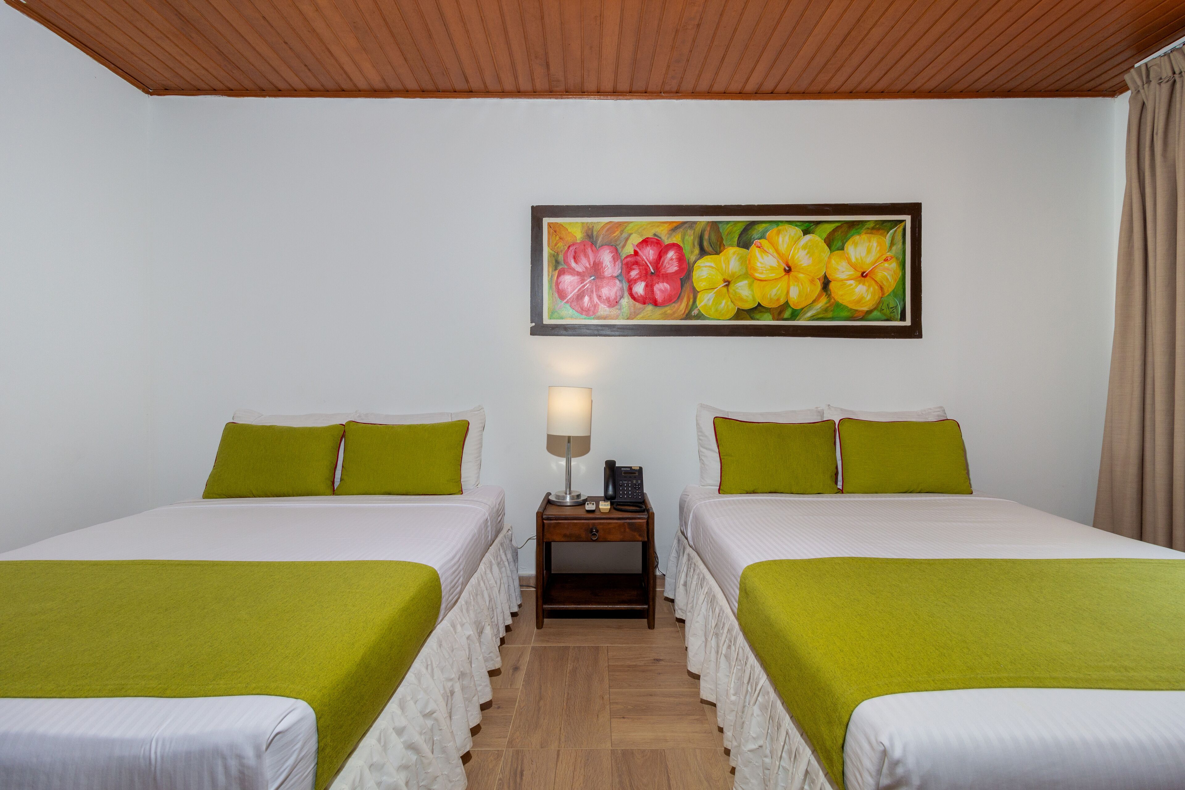 Une chambre d’hôtel avec deux lits, chacun recouvert d’une couvre-lit verte et doté de draps blancs. Une table de chevet avec une lampe et un téléphone se trouve entre les deux lits. Un tableau représentant des fleurs est accroché au mur, au-dessus des lits.