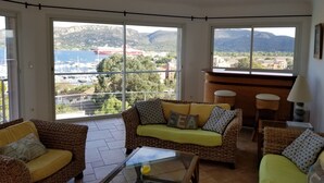Un salon meublé de meubles en osier, avec une table basse en verre et une vue sur un port de plaisance et des montagnes.