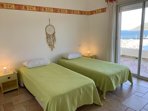 Quarto com duas camas, cada uma com colcha verde e almofadas brancas. Há um sonho preso na parede, uma mesa de cabeceira com abajur e uma janela com vista para o mar e barcos.