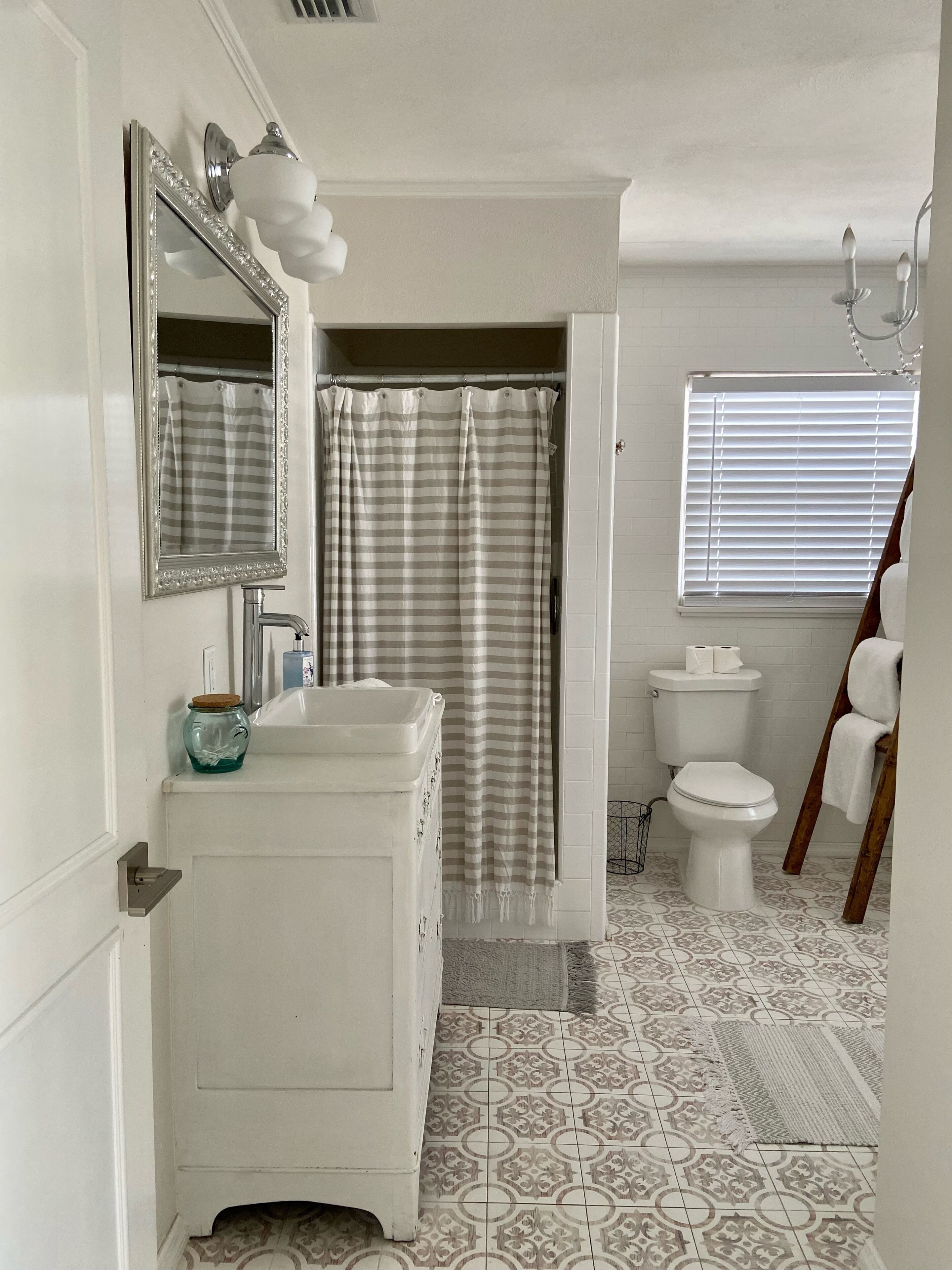 Un bagno con lavandino bianco, specchio e servizi igienici. Si vede una tenda da doccia a righe e una finestra con persiane.