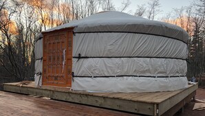 Una yurta con puerta de madera, rodeada por una terraza de madera, con árboles y un cielo al atardecer como telón de fondo.