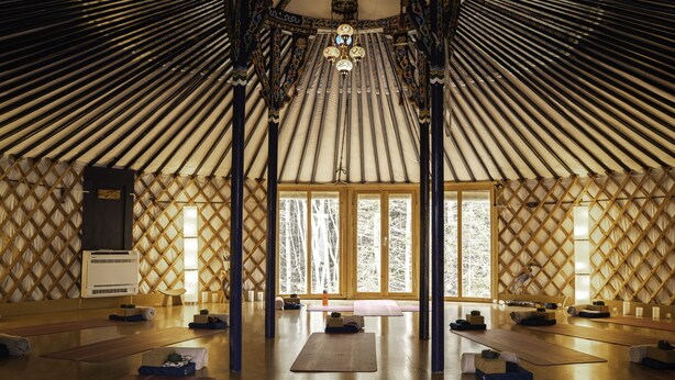 Un interior espacioso con techo alto y un área central abierta, presumiblemente un espacio para yoga o meditación, rodeado de bancos y colchonetas de madera.