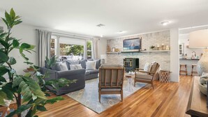 Un salon moderne avec un canapé gris, des chaises en bois, une cheminée et un coin cuisine visible à travers une porte ouverte.