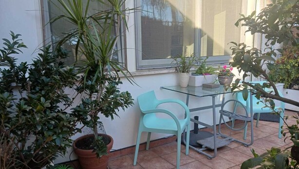A patio area with a glass table and two turquoise chairs, surrounded by potted plants and a window with blinds.
