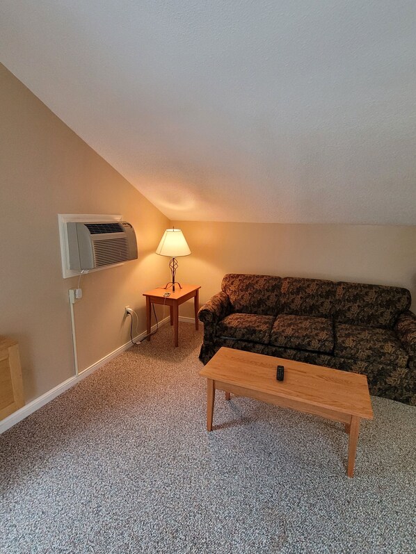 A small, carpeted room with a sofa, a wooden table, a lamp, and an air conditioning unit mounted on the wall. - Beautiful condo in Bartlett NH July 24th - July 31st 2027 PERFECT SUMMER WEEK (Bartlett)
