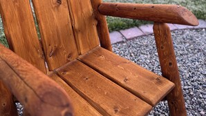 A wooden Adirondack chair with a metal frame, positioned on a gravel path with a grassy area and trees in the background.