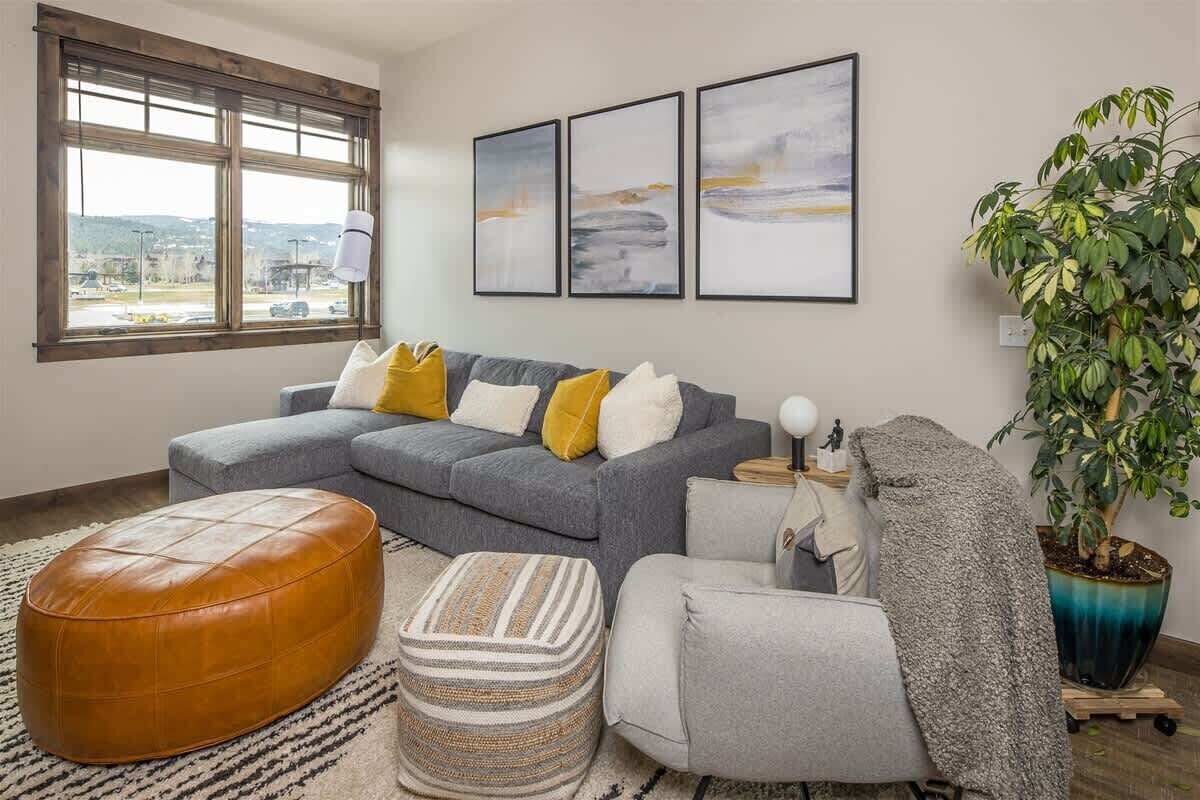 A modern living room with a grey sectional sofa, a brown leather ottoman, a grey armchair, and a large potted plant.