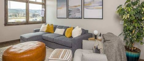 A modern living room with a grey sectional sofa, a brown leather ottoman, a grey armchair, and a large potted plant.