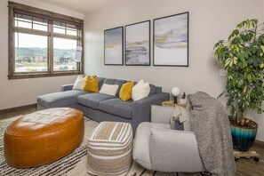A modern living room with a grey sectional sofa, a brown leather ottoman, a grey armchair, and a large potted plant. - Modern Luxury 1 Bedroom Condo Big Sky Town Center (Big Sky)