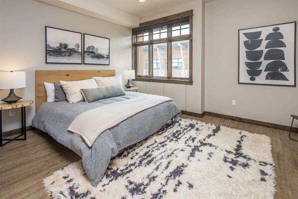 A bedroom with a large bed, two bedside tables with lamps, a window with a view, and artwork on the walls.