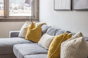 A grey sofa with white and yellow cushions, a floor lamp, and a window with a view of buildings.