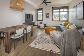 A modern living room with a wooden dining table, grey sofa, and a large window offering a view of the outdoors.