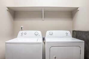 Two white front-loading washing machines in a laundry room with a white wall and a shelf above.