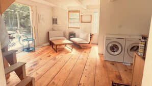 A rustic cabin interior with wooden floors, a sofa, a chair, a small table, a washing machine, and a staircase.