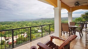 A balcony with a view of a green landscape, a table with chairs, and a ceiling fan. - Amazing Ocean View atop Coco Cliffs (Playa del Coco)