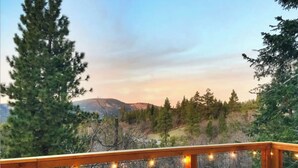A wooden deck with a railing, overlooking a mountainous landscape with trees and a clear sky.