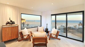 A modern living room with a large window overlooking a beach, wooden furniture, and a guitar. - Oceanfront Terrasol Condo with Patio Fireplace (Cabo San Lucas)