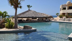 A clear pool with a thatched-roof structure, palm trees, and a building with balconies. - Oceanfront Terrasol Condo with Patio Fireplace (Cabo San Lucas)