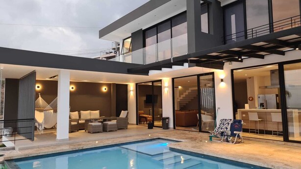 Ein modernes, zweistöckiges Haus mit Swimmingpool, Außenbereich mit Sitzgelegenheit und einer durch Glastüren einsehbarer Küche.