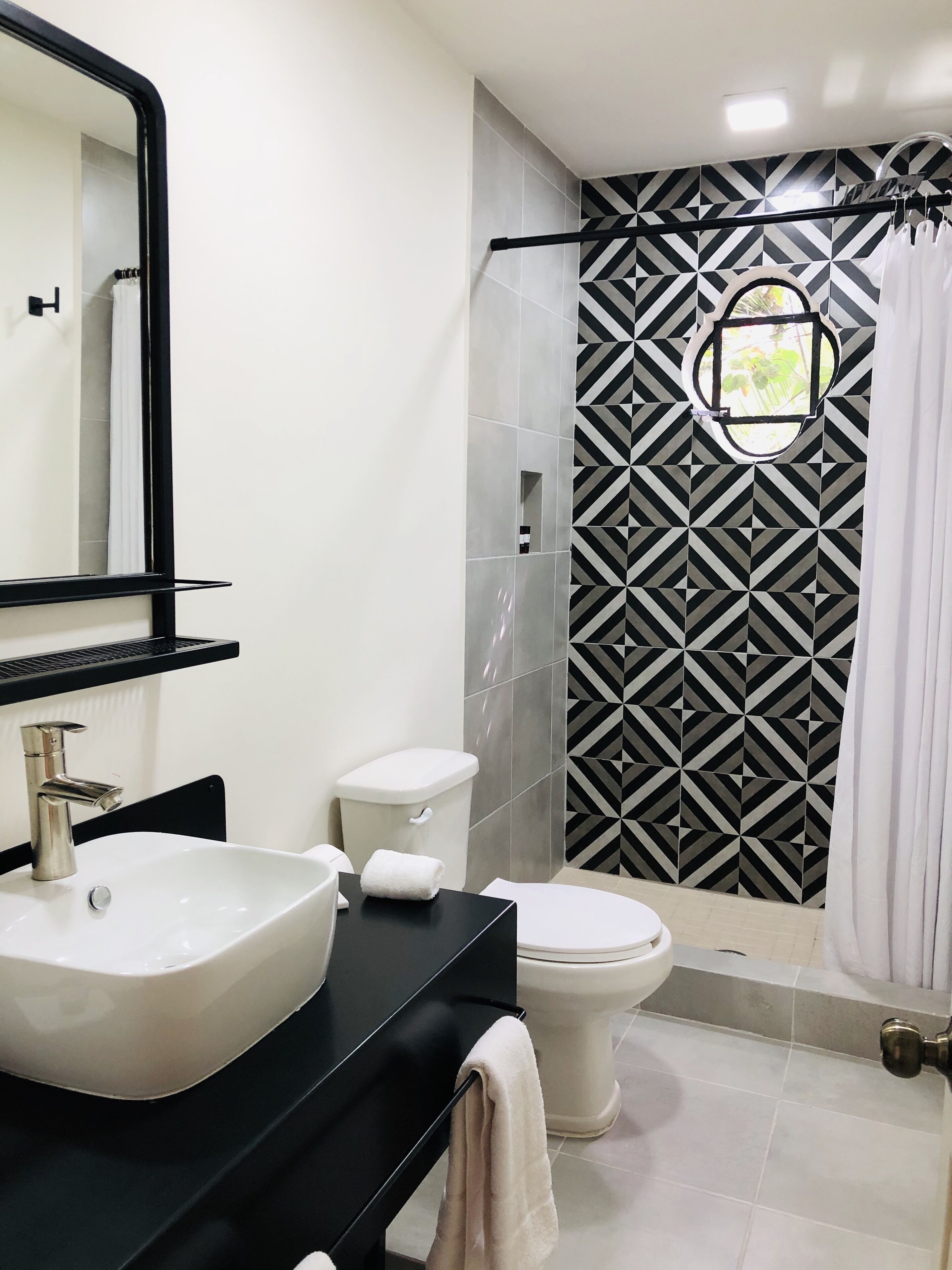A modern bathroom with a black and white geometric tile backsplash, a white sink, a black countertop, a white toilet, and a glass shower enclosure.