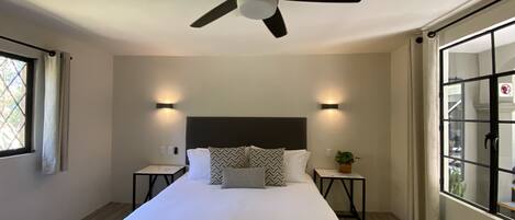 A hotel room with a single bed, a ceiling fan, two bedside tables with lamps, a window with curtains, and a view of the outside.