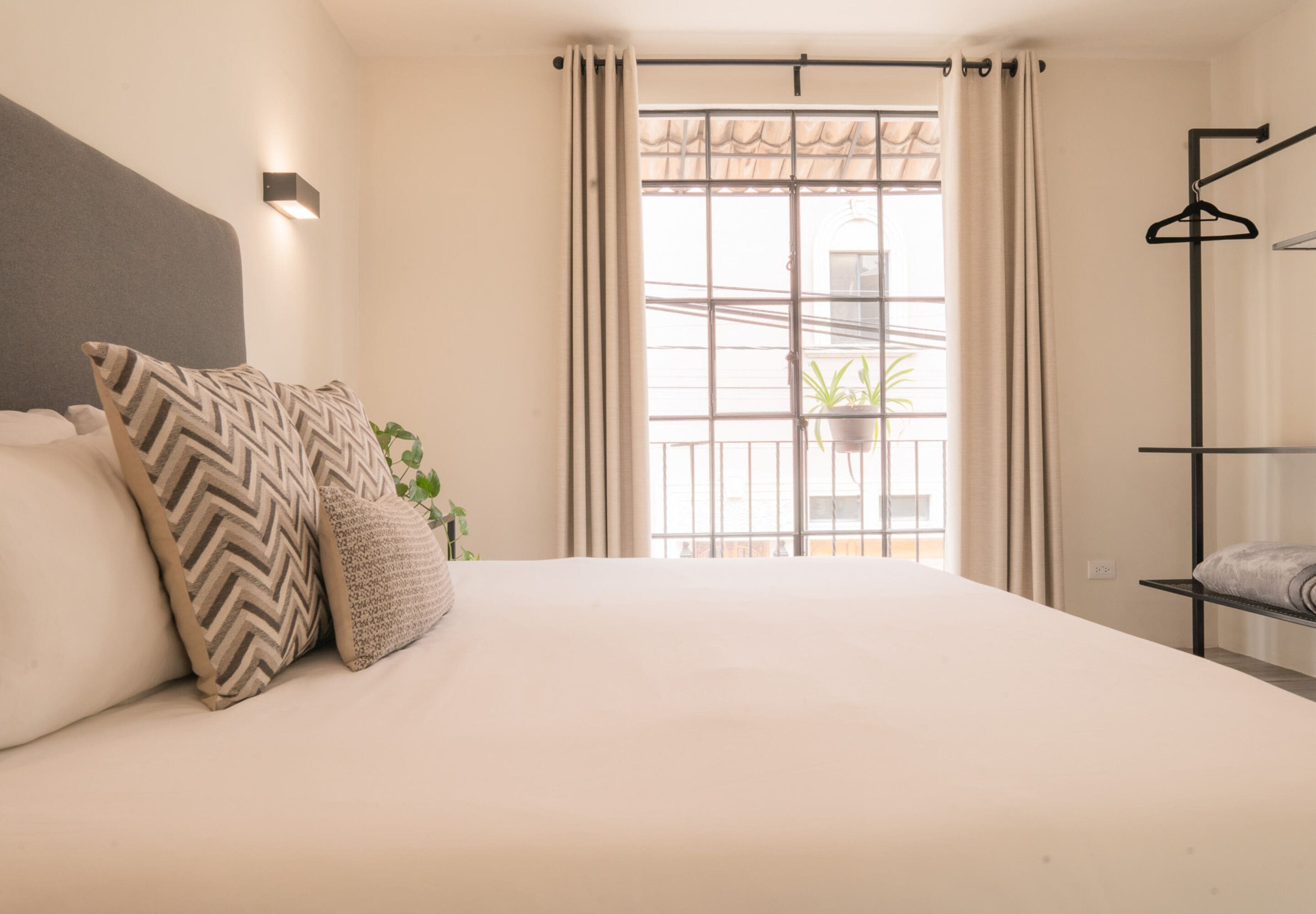 Une chambre à coucher avec un lit, une plante suspendue, une fenêtre avec des rideaux et un luminaire fixé au mur.