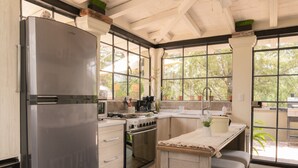 A modern kitchen with a stainless steel refrigerator, wooden cabinets, a built-in oven, and a large window offering a view of greenery. - Casa Doper Guadiana (San Miguel de Allende)
