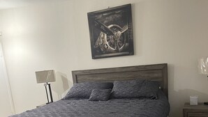 Et soveværelse med en træseng, et mørkt mønstret sengetæppe, et ophængt kunstværk, et natbord med en lampe og et sengebord med en tissueske.