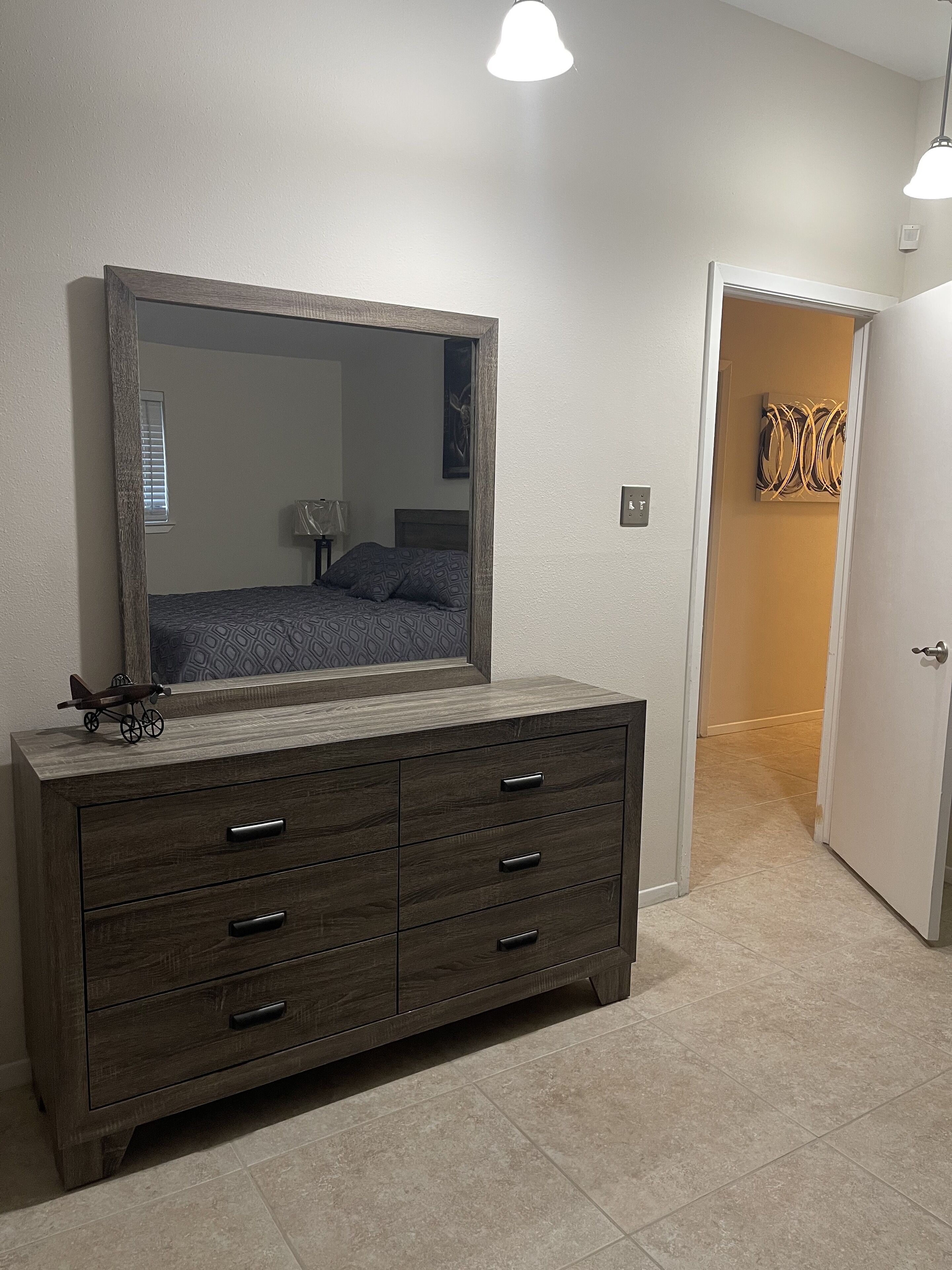 A bedroom with a large mirror, a dresser with drawers, a bed with patterned bedding, and a door leading to another room.