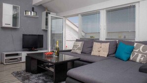 Ein modernes Wohnzimmer mit grauem Sofa, einem schwarzen Couchtisch, einem Flachbildfernseher und einem großen Fenster mit Jalousien.